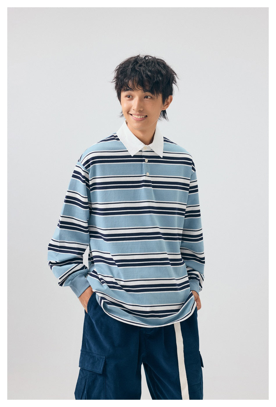 Smiling model wearing a light blue, navy and white striped rugby shirt with a white collar and navy trousers.