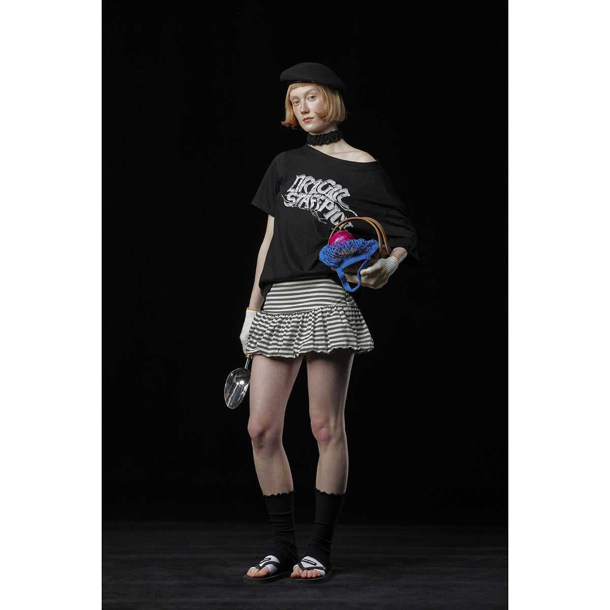 Model wearing a black graphic T-shirt with a gray-and-black striped ruffle skirt, beret and sandals.