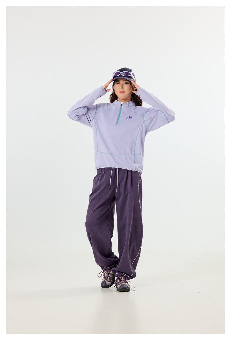 Full-length view of a woman wearing a lavender quarter-zip pullover with teal seam accents and dark purple wide-leg pants.