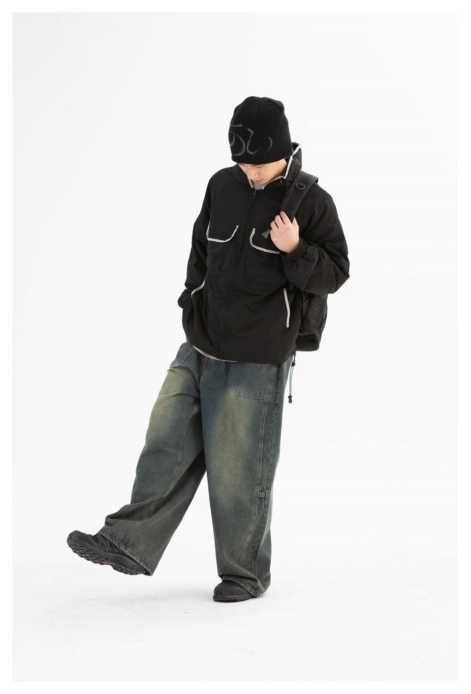 Man in a black hooded jacket and loose jeans, lifting one leg in a studio pose.