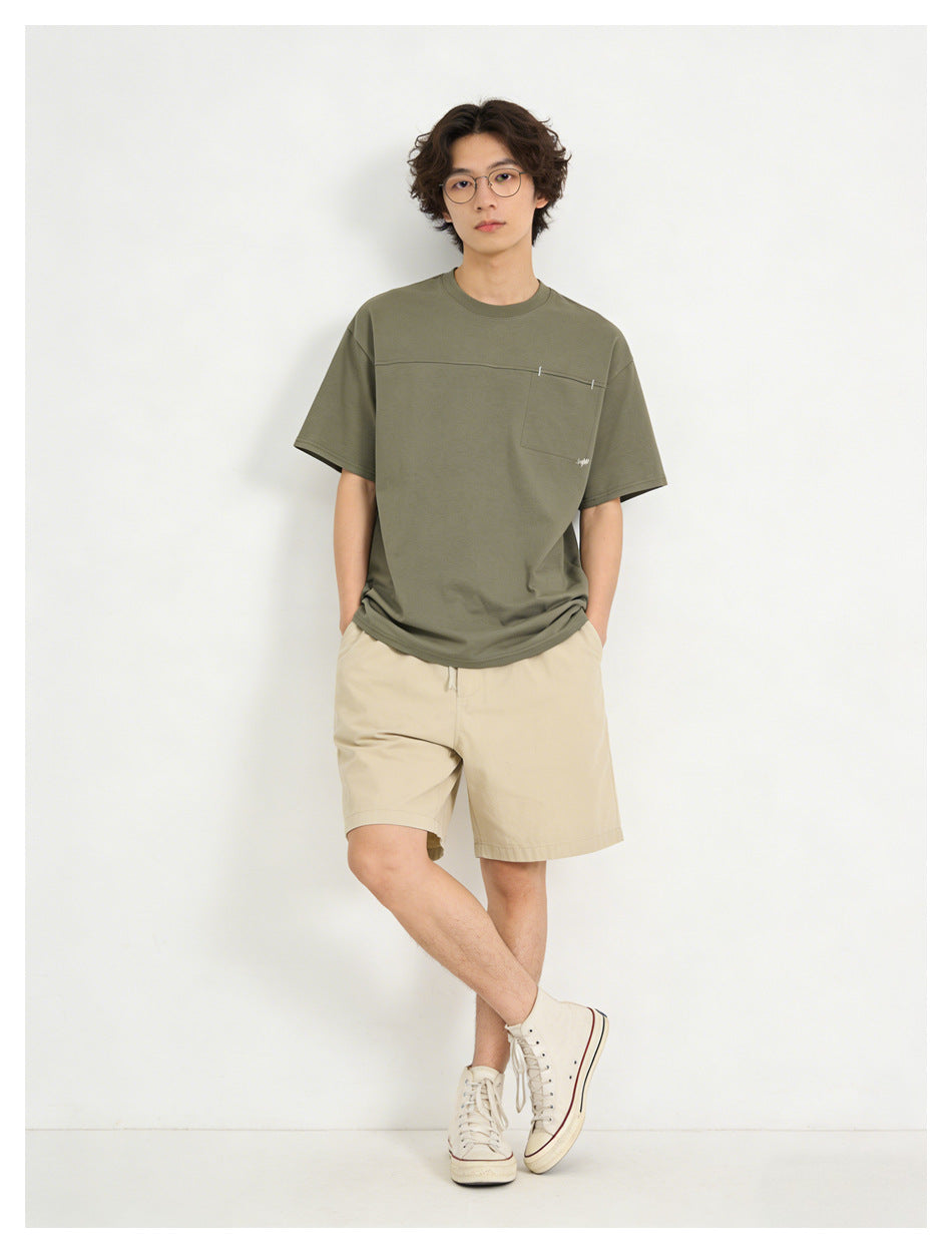 Model wearing an olive short-sleeve pocket T-shirt, beige shorts, and white high-top sneakers.