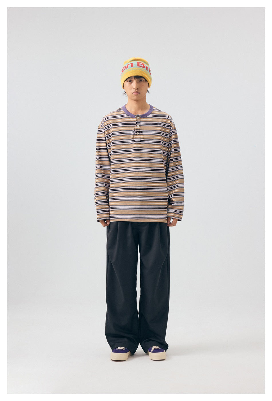 Full-length front view of a model in a tan striped henley, black wide-leg trousers, and a yellow beanie.
