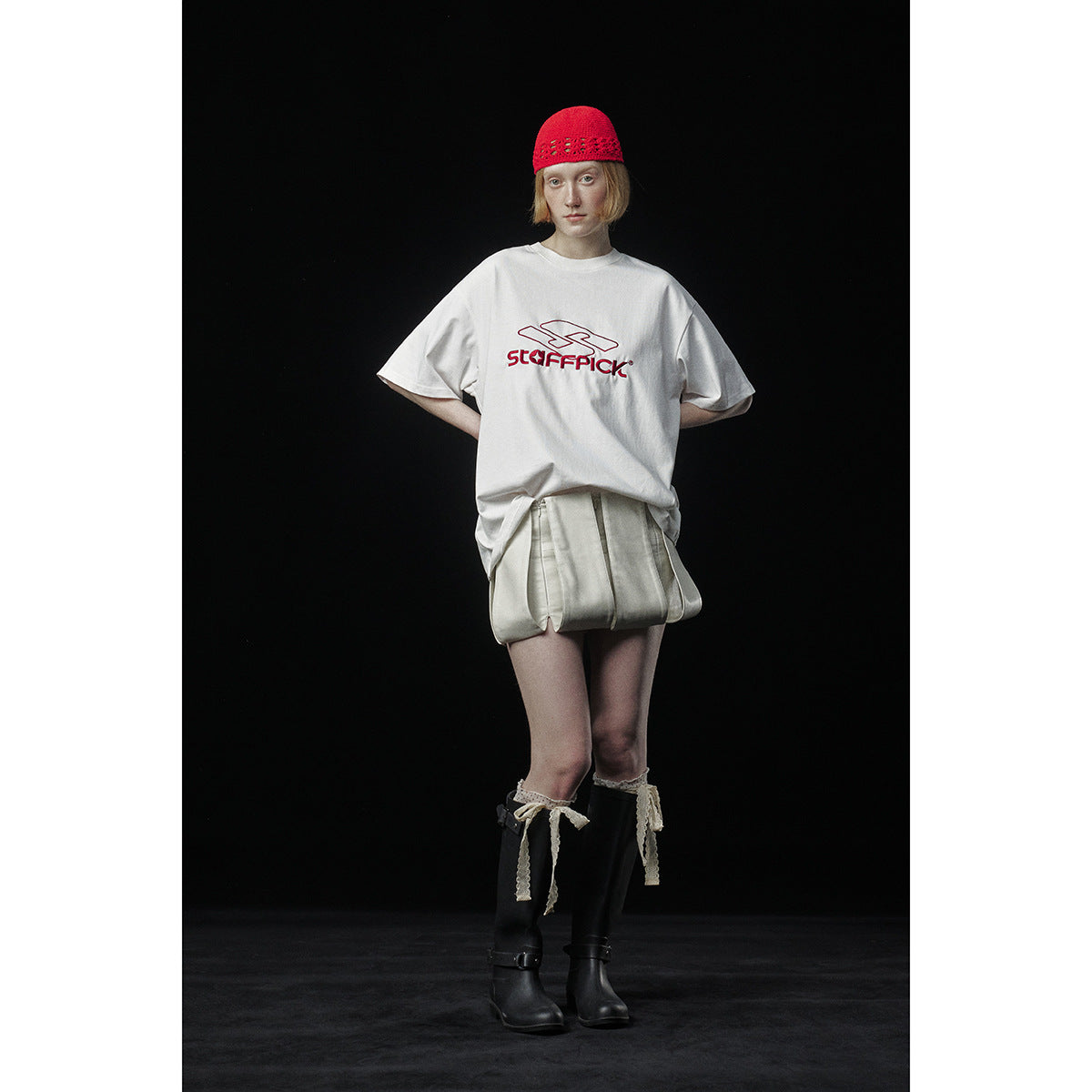 Model wearing an oversized white graphic T-shirt and pale shorts, with a red knit cap and black boots.