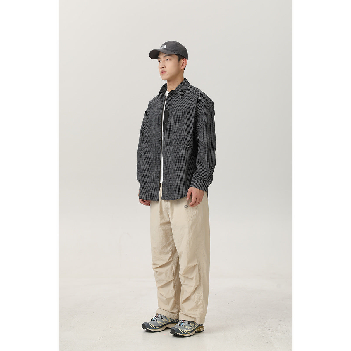 Full-length model wearing a charcoal micro-check button-up shirt with a chest pocket and loose beige trousers.