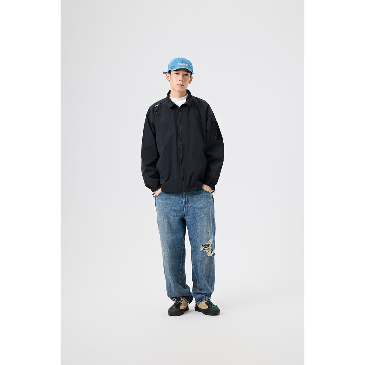Model wearing a black zip-front jacket with an angled chest zip and sleeve pocket, blue jeans, and a blue cap.