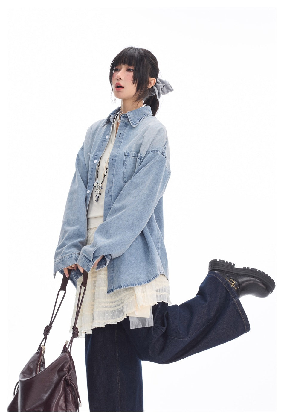 Person in a loose light blue denim shirt over a cream top and lace skirt, holding a bag with one leg raised