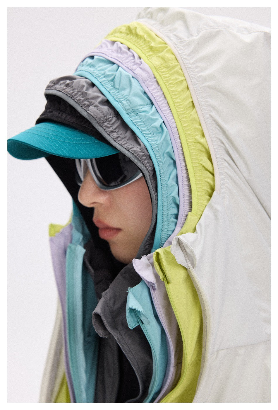 Close-up of layered jacket hoods in white, lime, lavender, aqua, and charcoal over a teal cap.