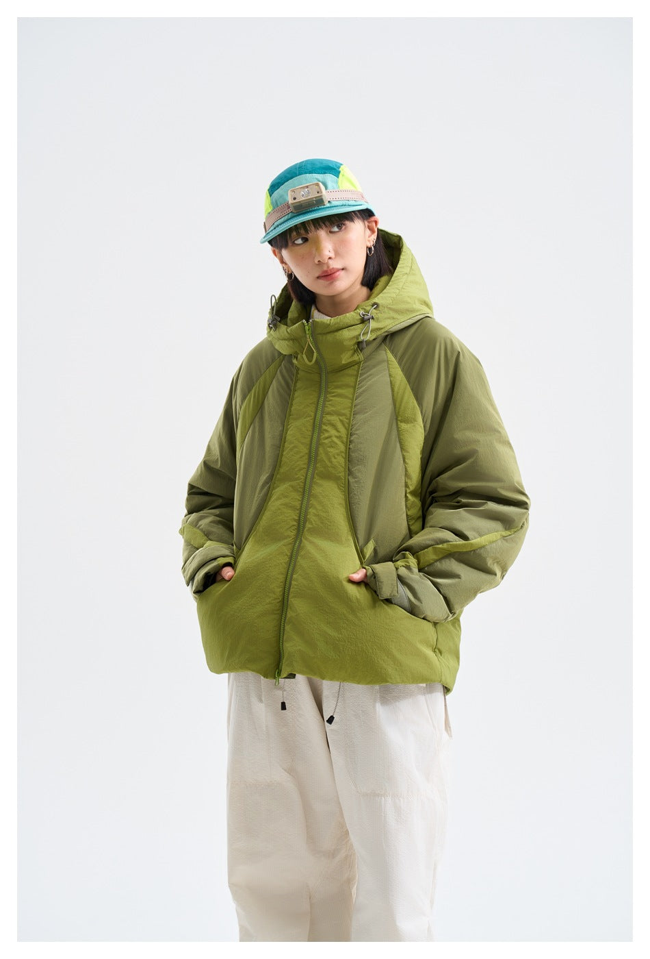 Model wearing an olive and lime-green color-blocked hooded jacket, white trousers, and a teal cap.