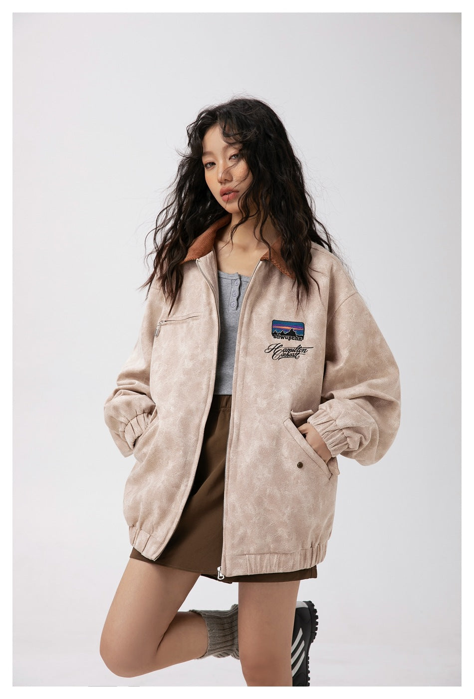 Model wearing a light beige zip jacket with a brown collar, chest embroidery, and elastic cuffs.