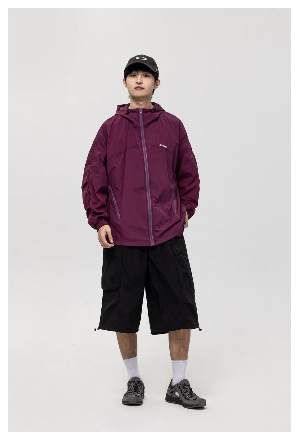 Full-length front view of a model in a burgundy hooded jacket, black shorts, and a black cap.
