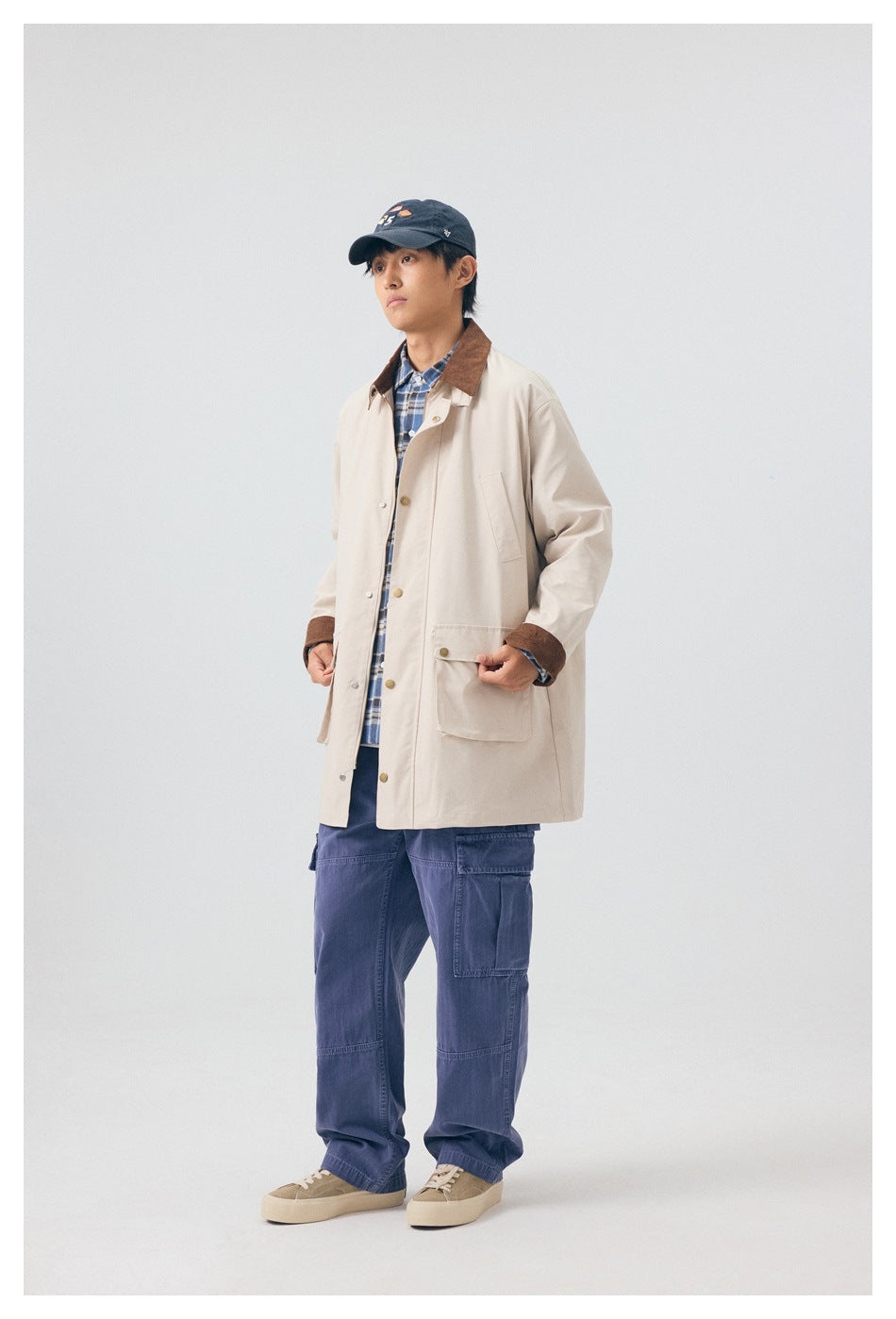 Model wearing a light beige coat with a brown collar and cuffs, blue plaid shirt, navy cargo pants, and cap.