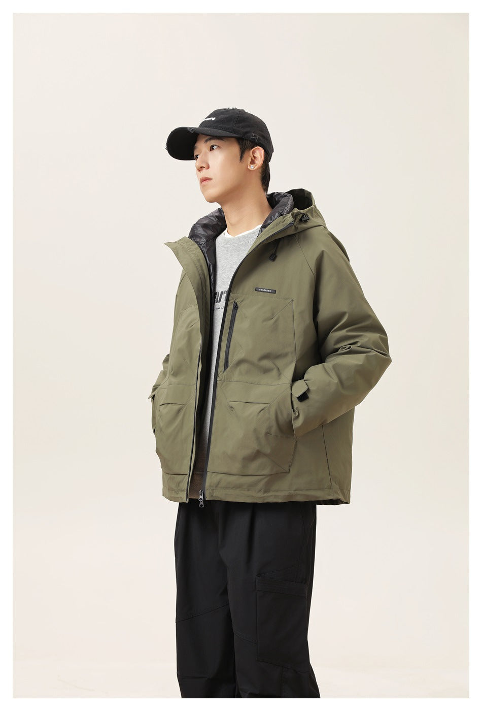 Model in army green hooded jacket over a gray sweatshirt with black pants, seen from the side with a hand in the pocket