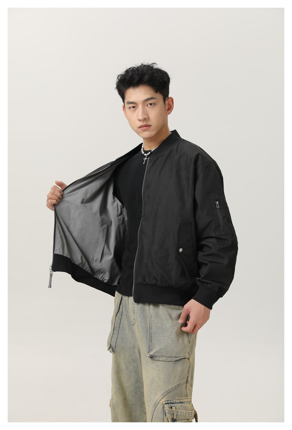 Man holding open a black bomber jacket to show the gray reverse side, with sleeve zip pocket and front snap pocket