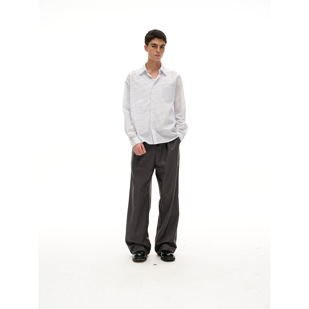 Full-length front view of a model in a white dashed-stripe button-up shirt, loose charcoal trousers and black shoes.