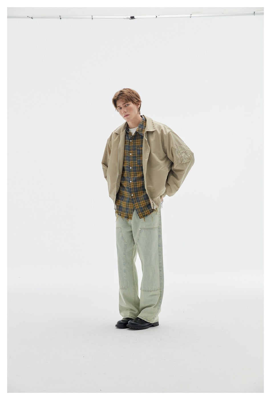Full-length model wearing a beige bomber jacket over a plaid shirt with loose pale green jeans and black shoes.