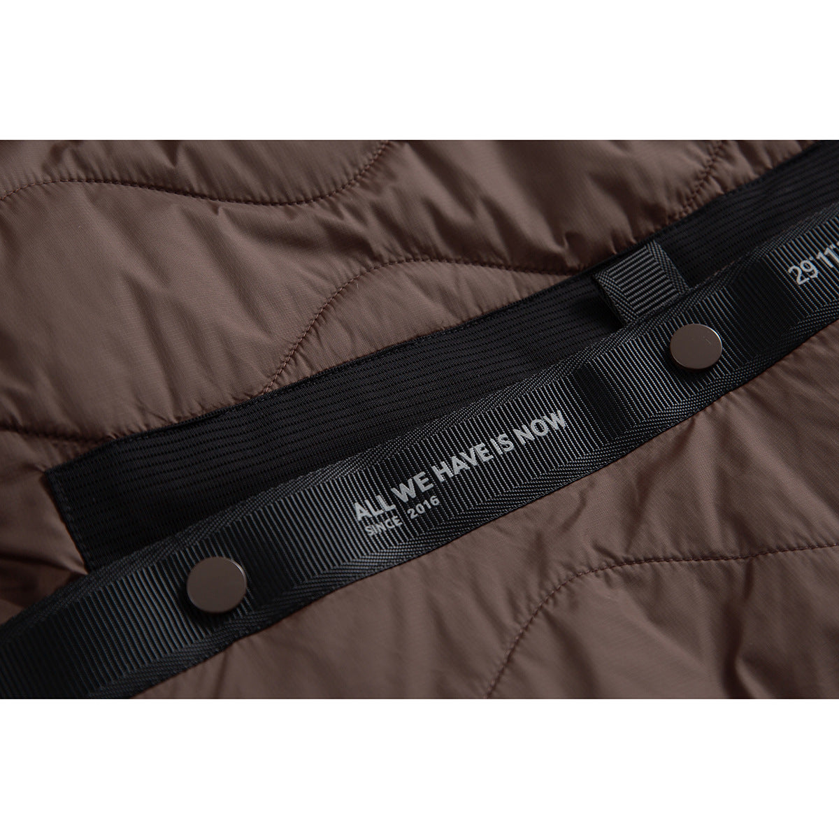 Close-up of the brown quilted jacket's black waist strap, snap fasteners and printed English text.