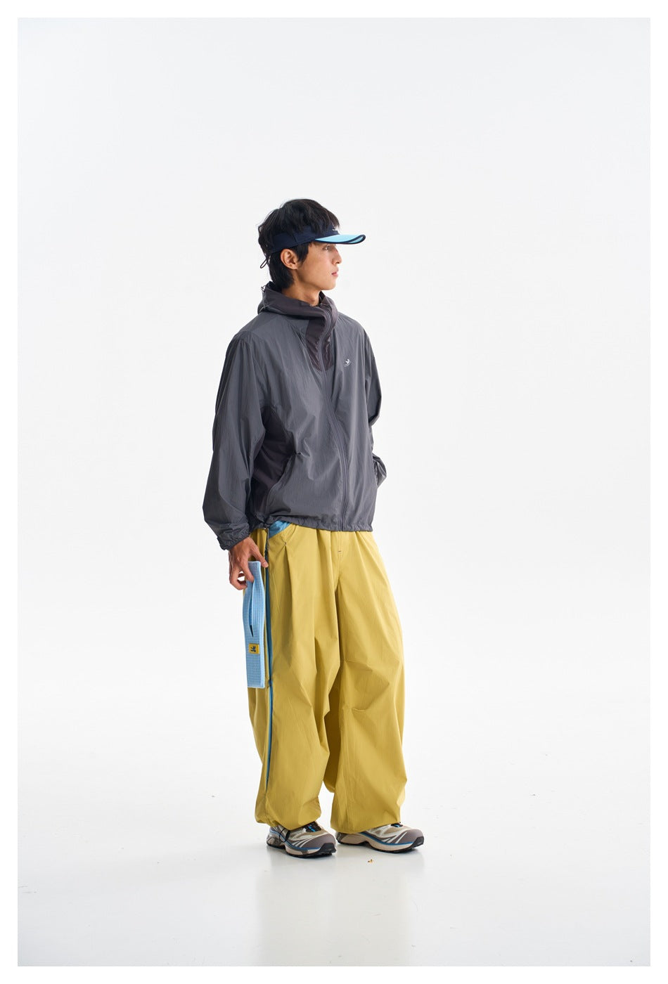 Three-quarter view of a person wearing a charcoal hooded jacket and loose mustard-yellow pants with blue side panels.