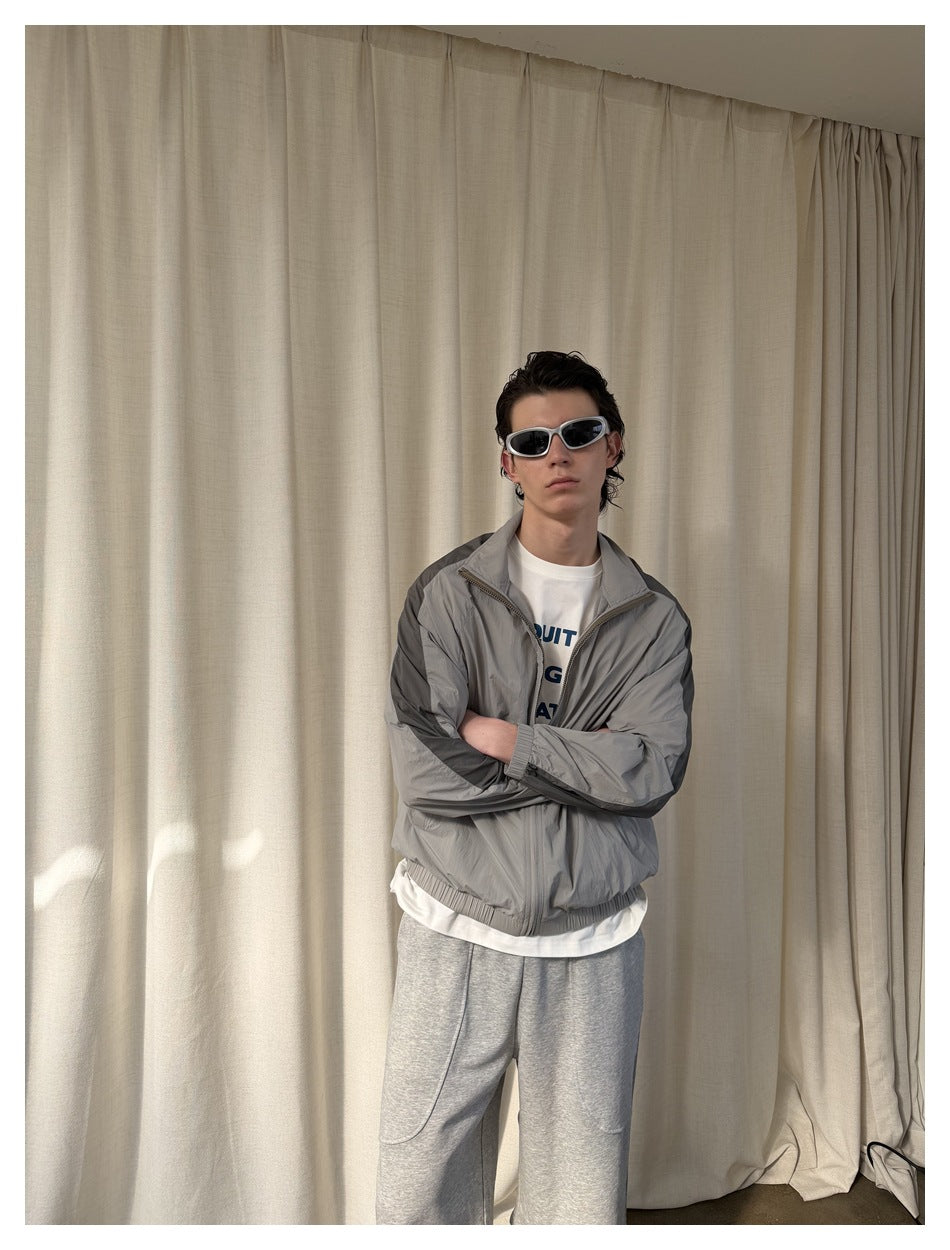 Model wearing a light gray zip-up jacket with dark shoulder stripes, shown from the front with arms crossed.
