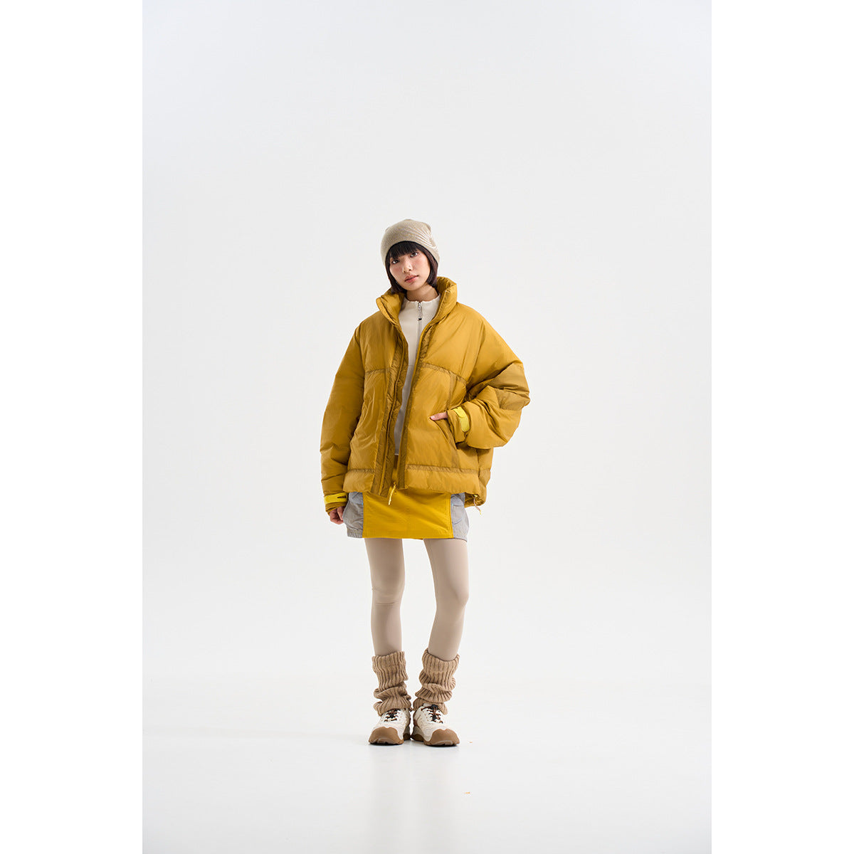 Model wearing a mustard-yellow oversized puffer jacket with a high collar, styled with a beanie and neutral layers.