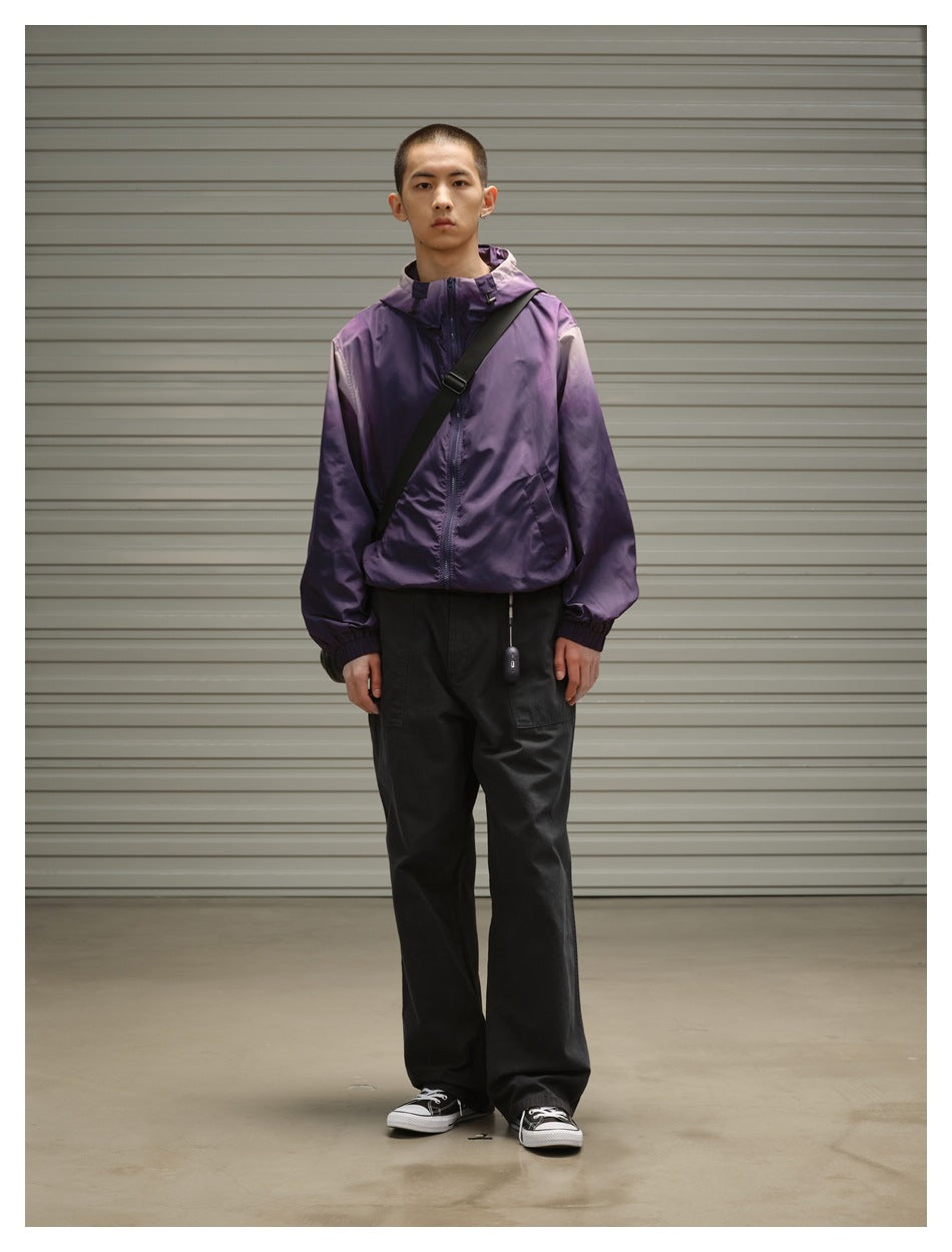 Full-length view of a man wearing a purple hooded zip jacket, loose black trousers and a black crossbody bag.