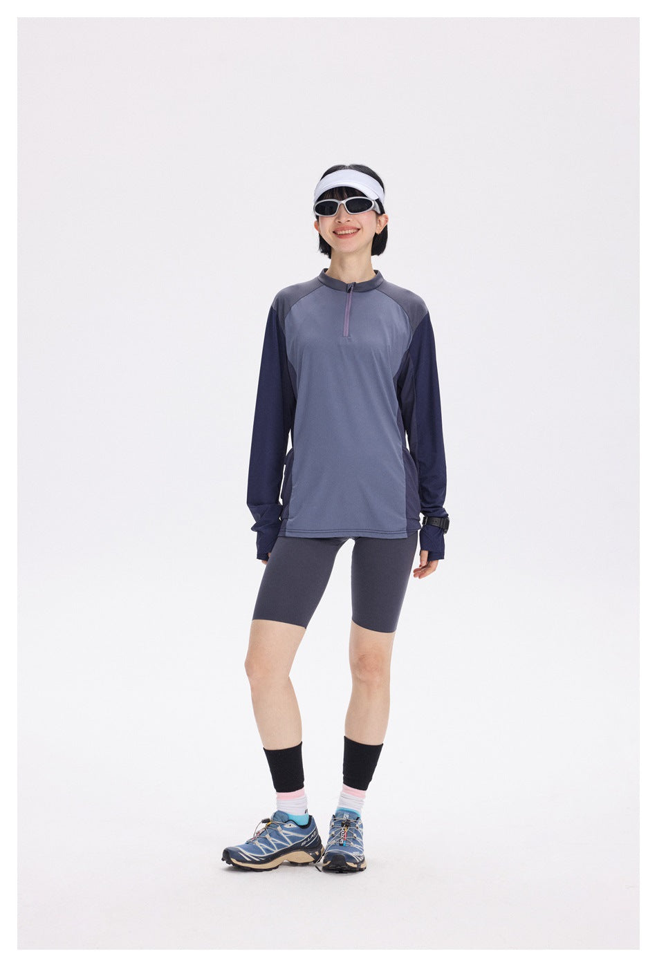 Full-length front view of a woman wearing a slate blue and navy quarter-zip top with dark cycling shorts.