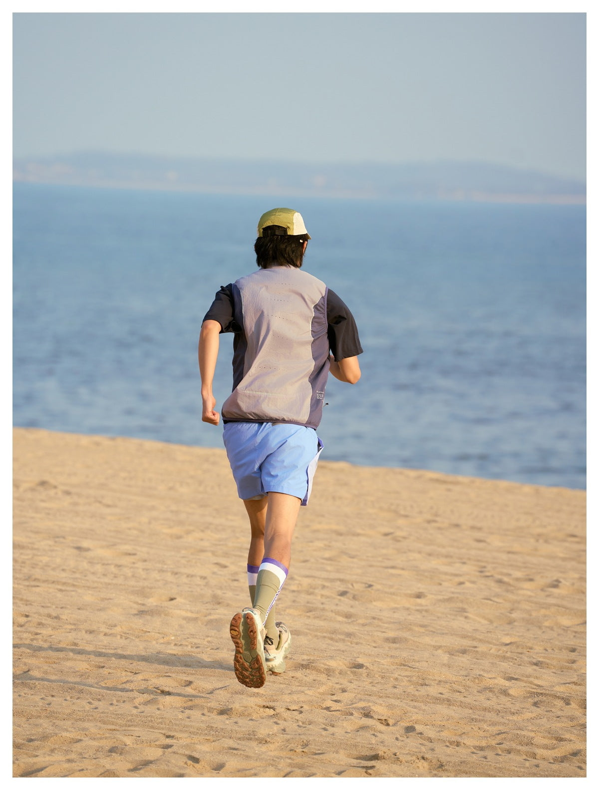 Rear view of a model jogging on a beach in a light gray-lavender sleeveless vest with dark side panels.