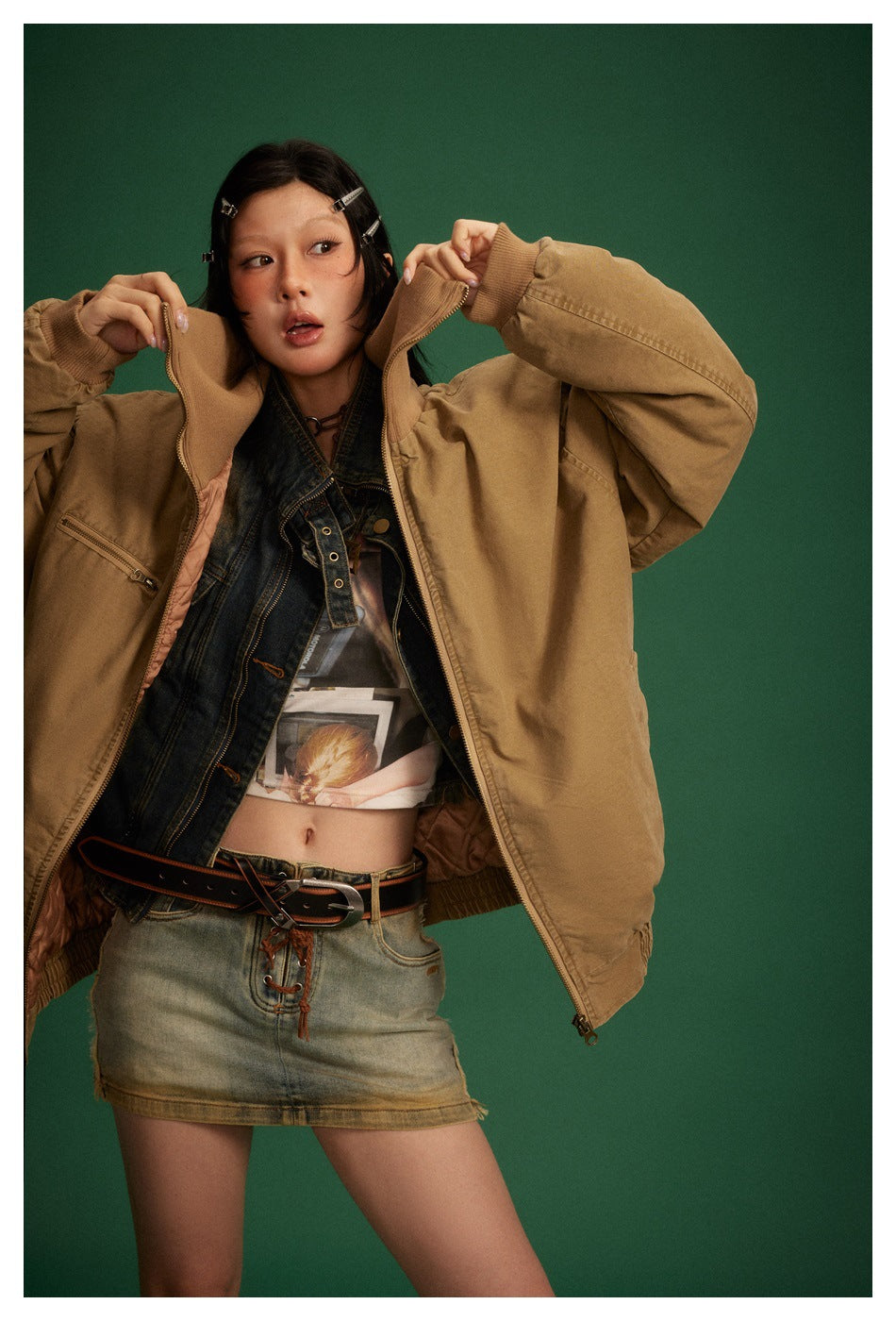 Model holds open the collar of an oversized camel-brown zip jacket, showing its ribbed cuffs and pink-toned lining.