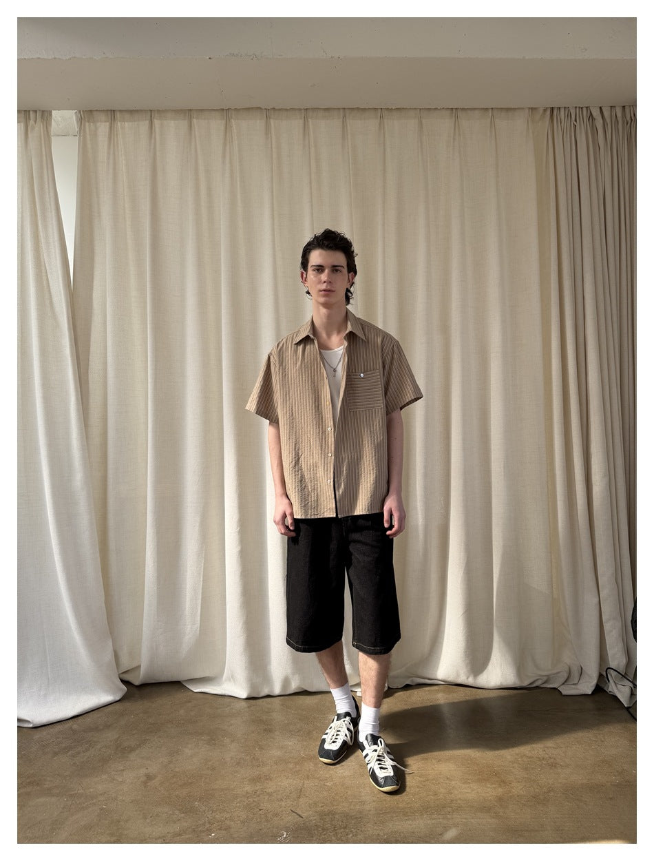 Man wearing khaki striped short-sleeve shirt open over white tee with black denim shorts, front view