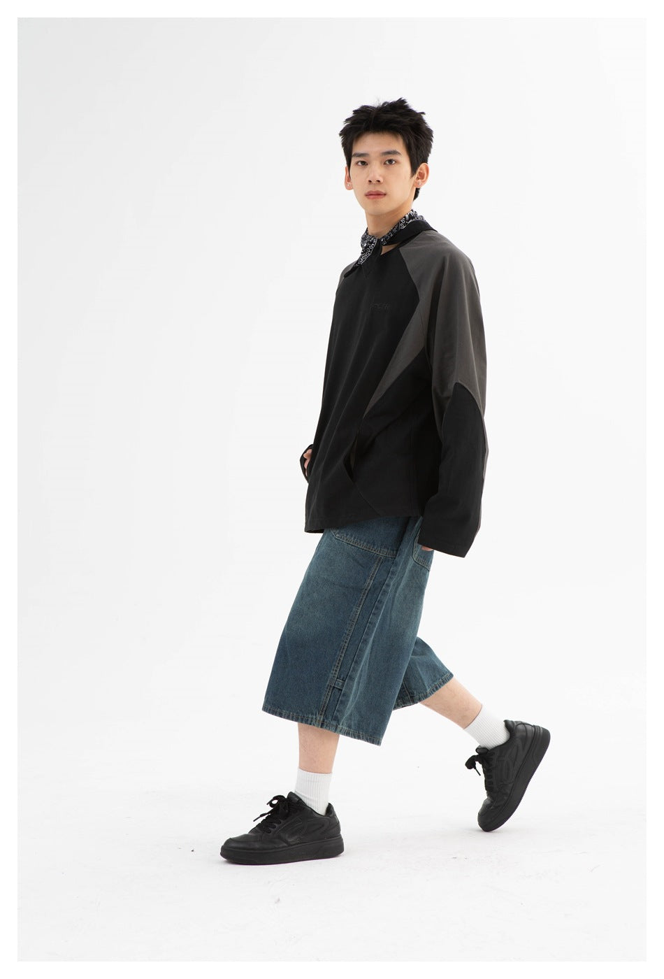 Angled view of a model wearing a black sweatshirt with charcoal panels and loose blue denim shorts.