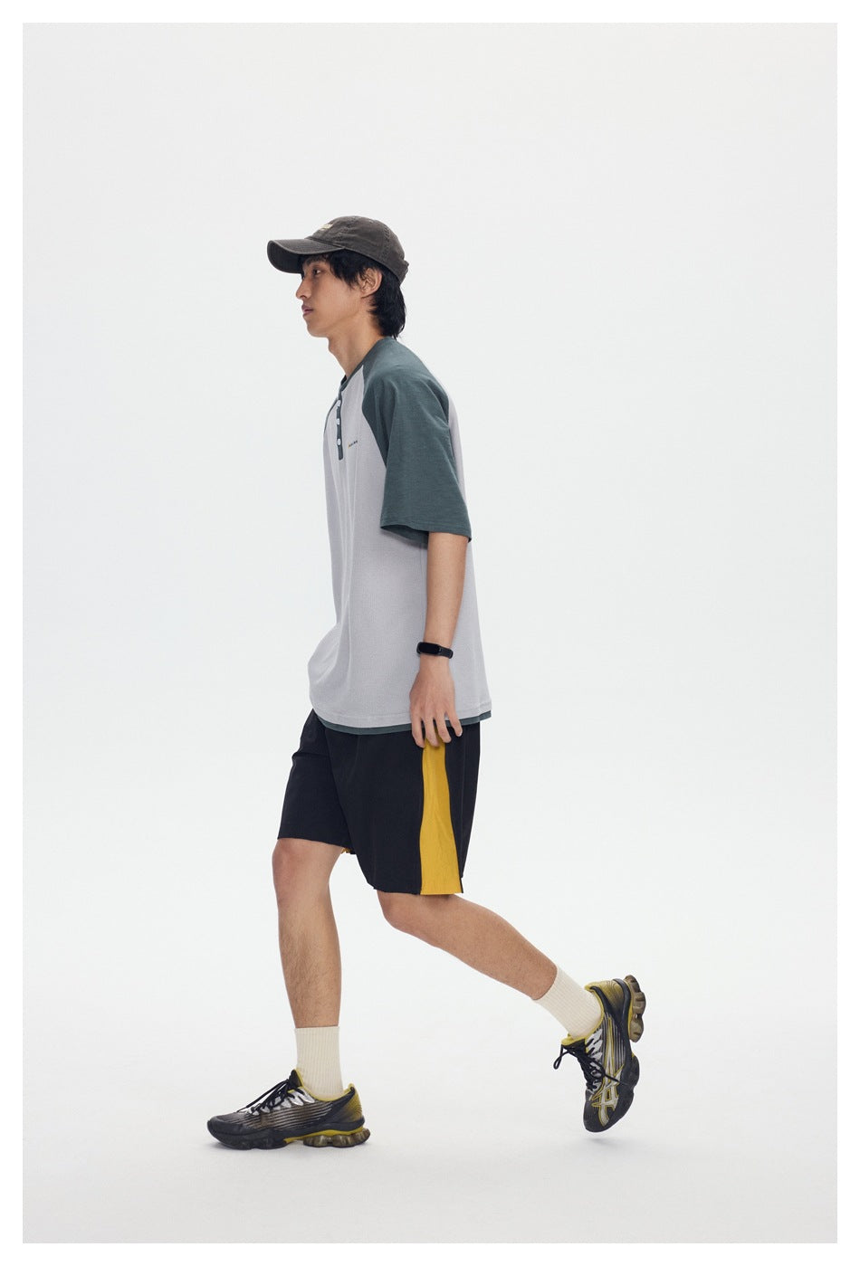 Side view of a model wearing a light gray raglan T-shirt with dark green sleeves and black shorts with yellow side panels.