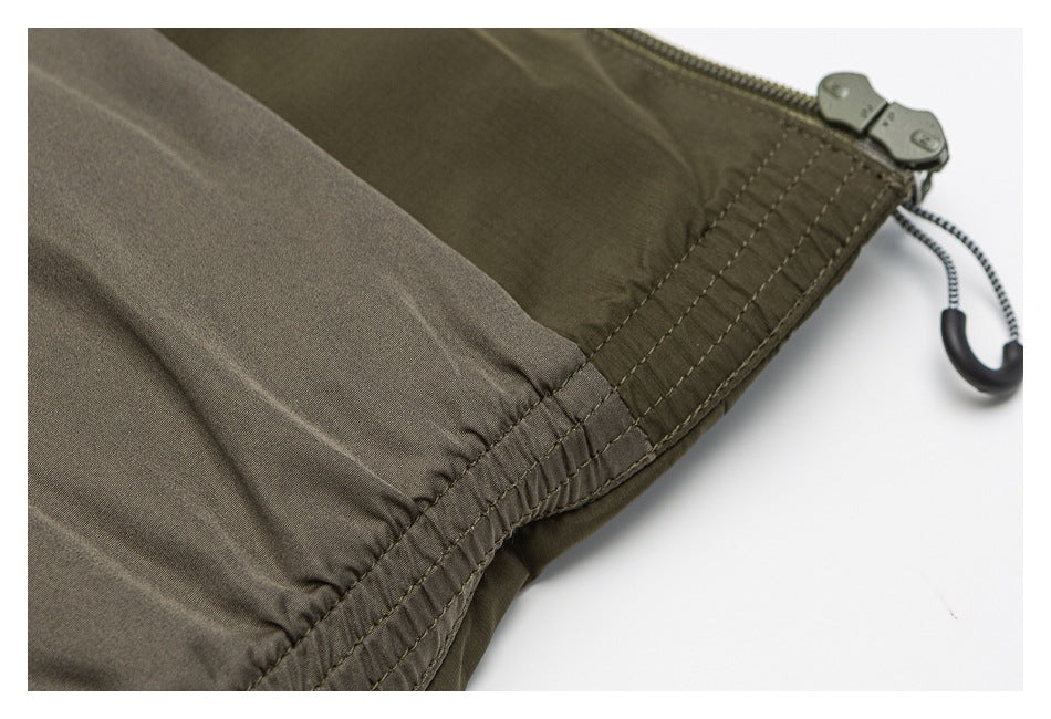 Close-up of an olive garment hem with gathered stitching, a zipper, and a hanging cord toggle.