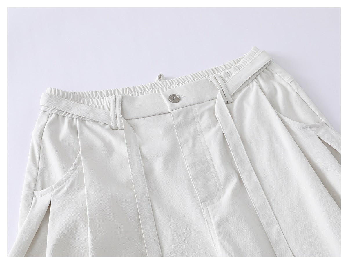 Close-up of ivory shorts showing an elasticated waistband, button, belt loops, front pleats, pockets and long belt tabs.