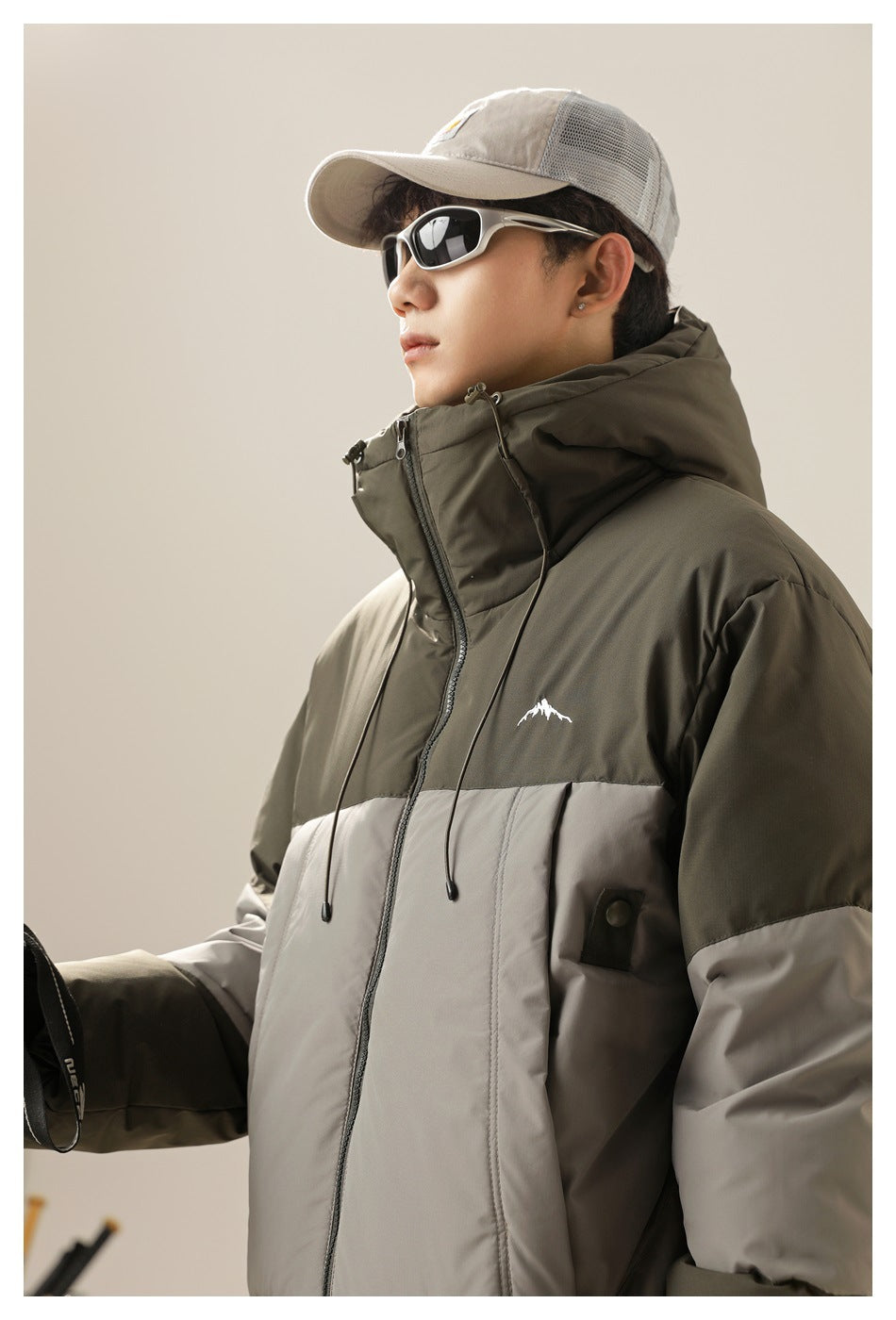 Close-up of a model wearing a dark olive and light gray color-block hooded jacket with a high collar and chest graphic.