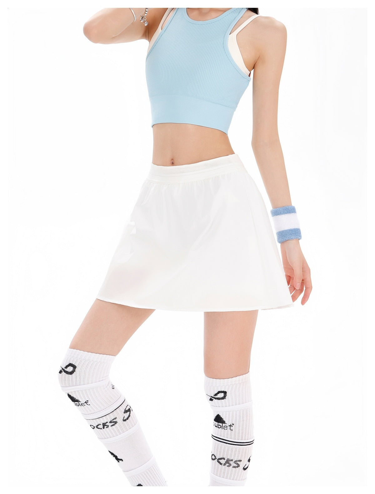 Model wearing a white tennis skirt with a white waistband and pale blue sleeveless crop top, front view.