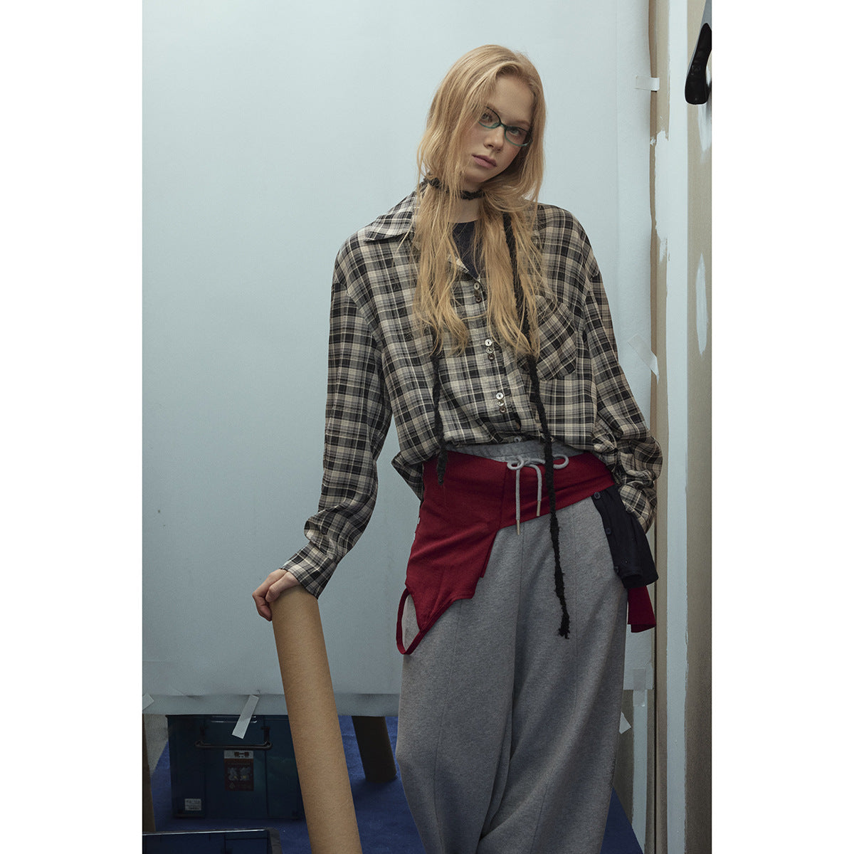 Model wearing a gray plaid button-up over layered tops, wide gray trousers and a red-and-black waist wrap.