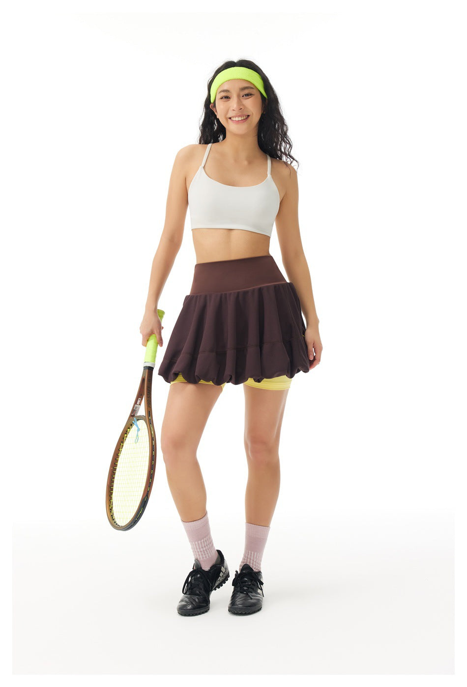 Front view of a model wearing a dark brown bubble tennis skirt with yellow built-in shorts, holding a tennis racket.