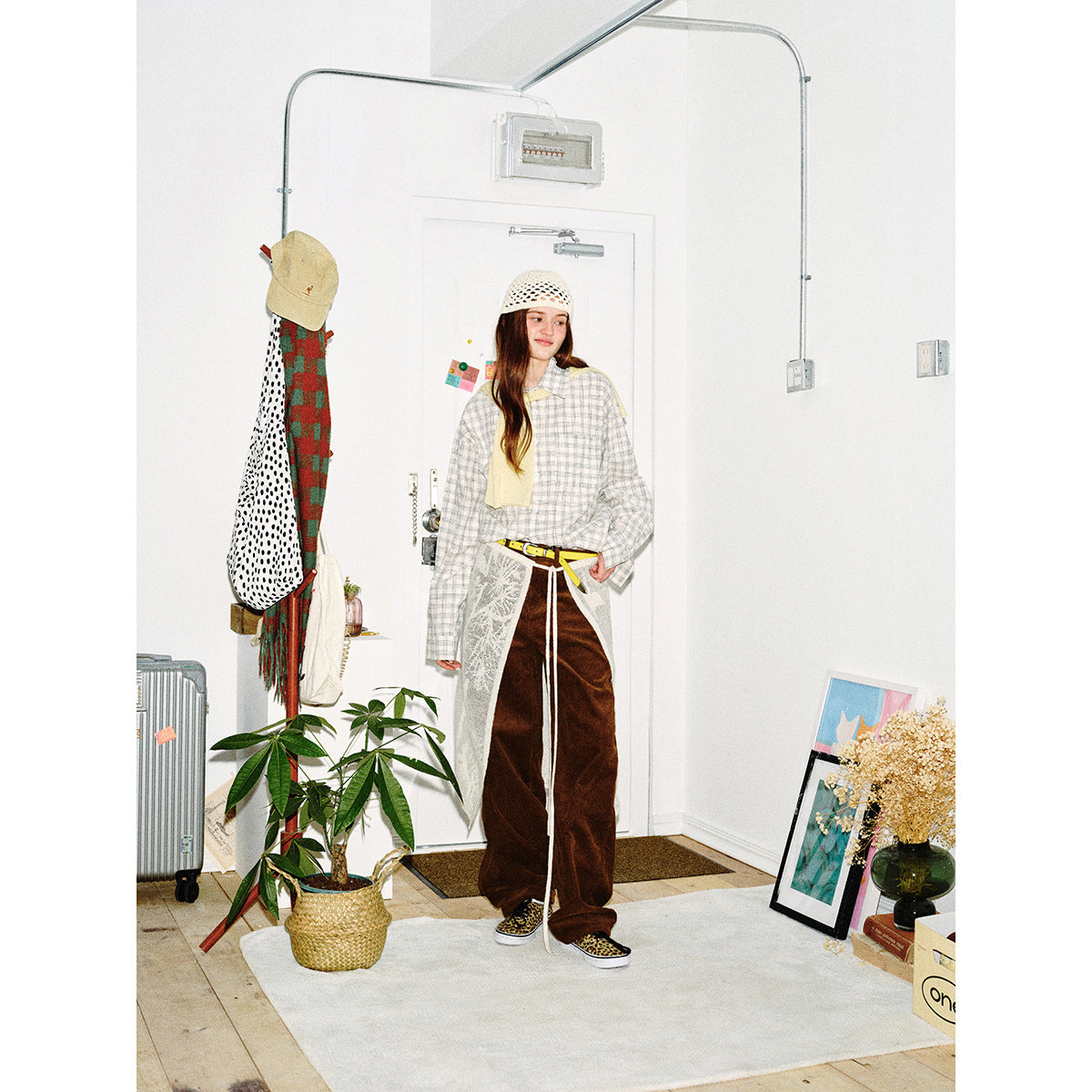Full-length front view of a model wearing a light plaid shirt, brown wide-leg pants, a white lace apron and crochet cap.