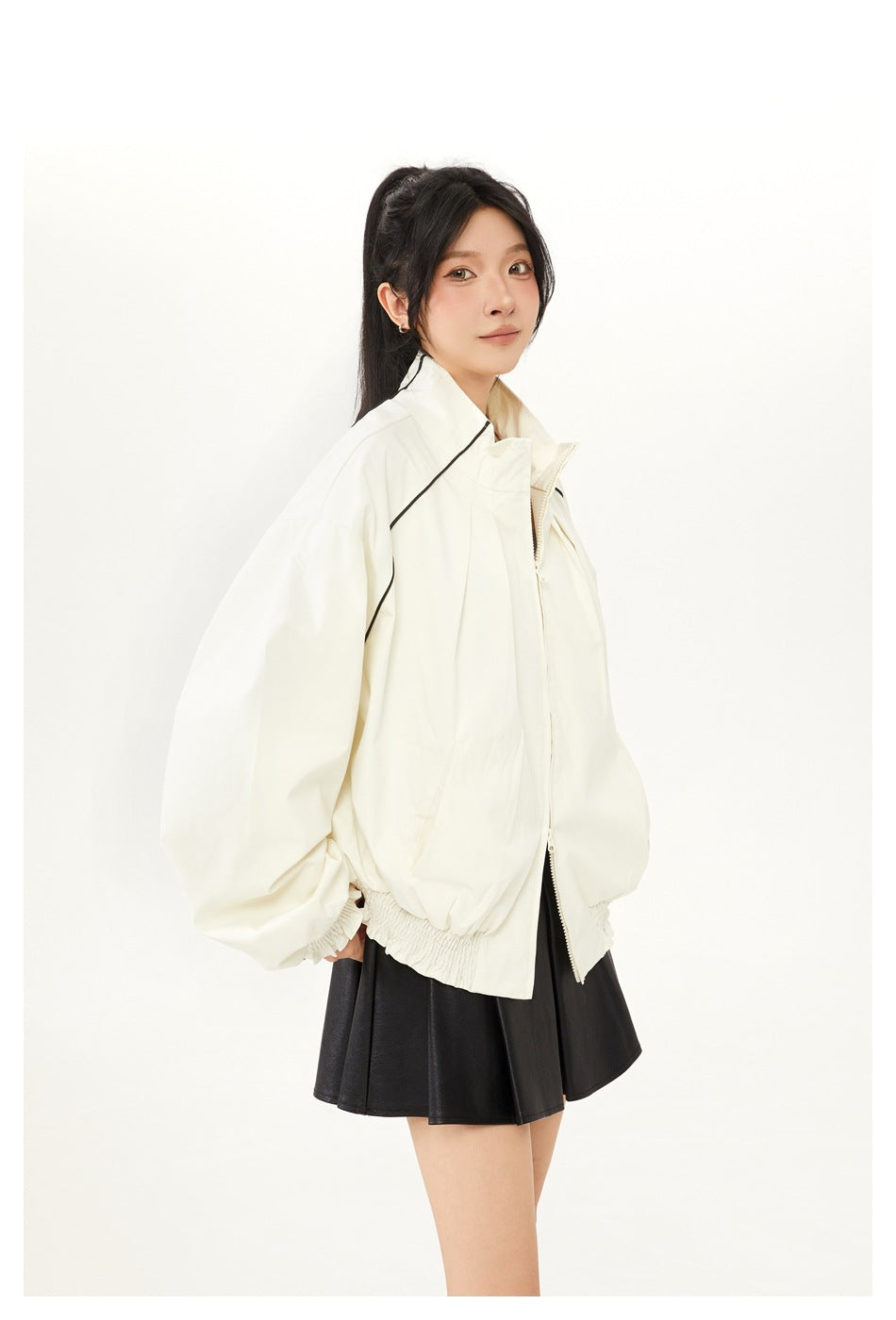 Model wearing an ivory zip-up jacket with a high collar, black piping, voluminous sleeves and a gathered hem.