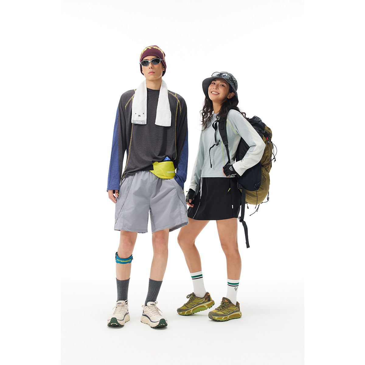 Two hikers pose in color-block long-sleeve tops, gray shorts and a black skirt, with hats and backpacks.