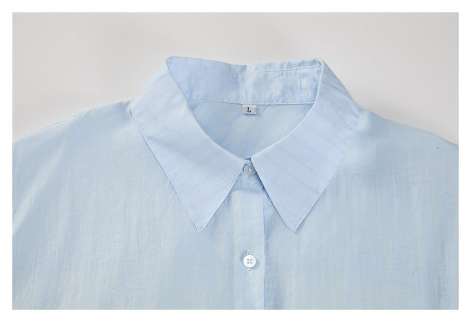 Upper front of a pale blue striped button-up shirt with a pointed collar and white buttons, laid flat.