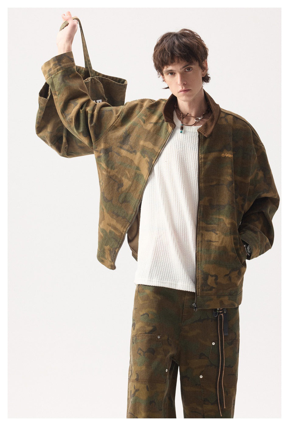 Model wearing a brown-green camouflage jacket and matching trousers, holding up a matching tote by its strap.
