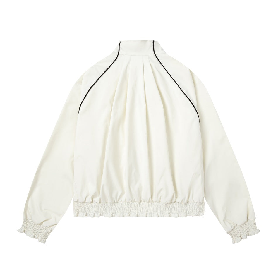 Back view of an ivory jacket laid flat, with black curved piping, a pleated back, and gathered cuffs and hem.