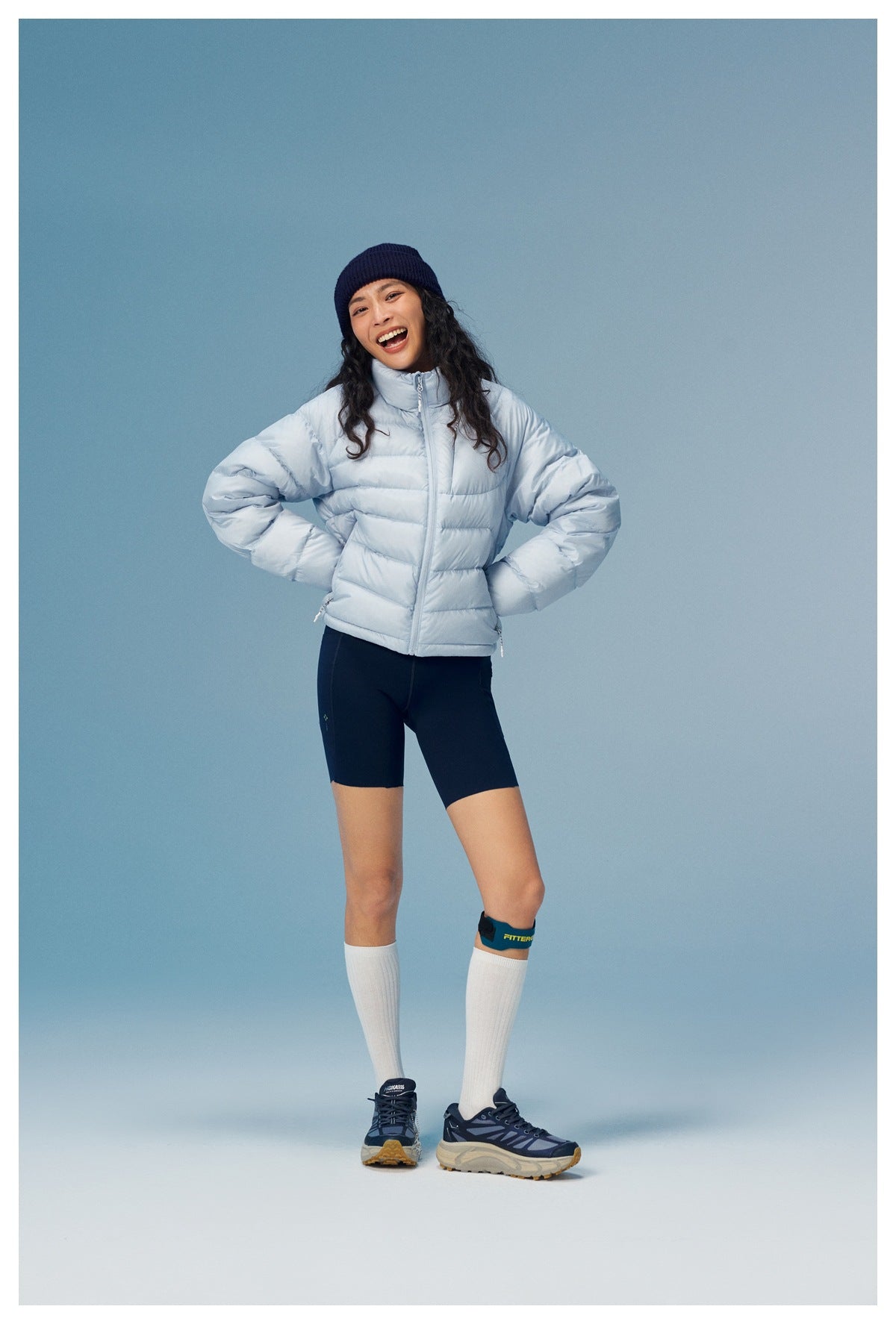 Model wearing a light blue puffer jacket, navy shorts and a dark knit beanie, shown from the front.