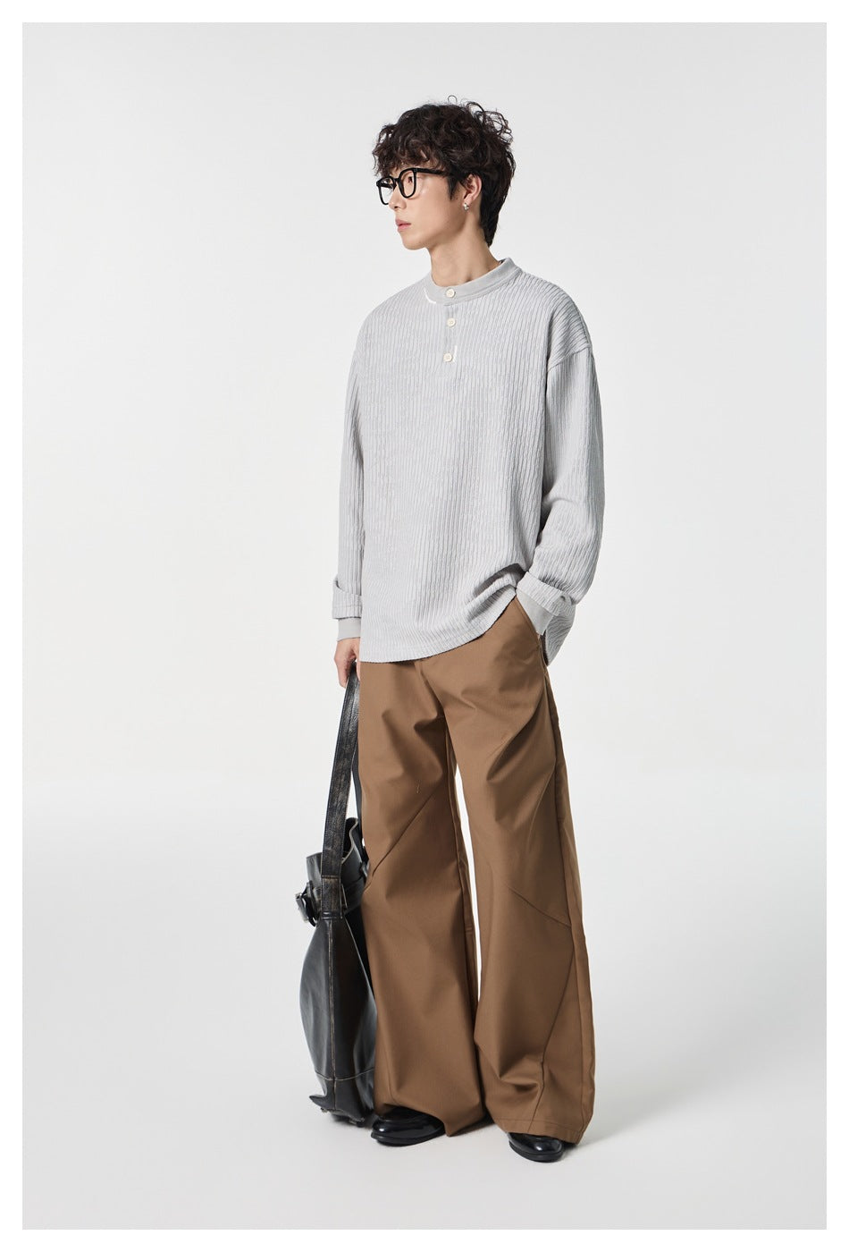 Side view of a model wearing a pale gray textured button-neck top and wide brown trousers, carrying a black bag.