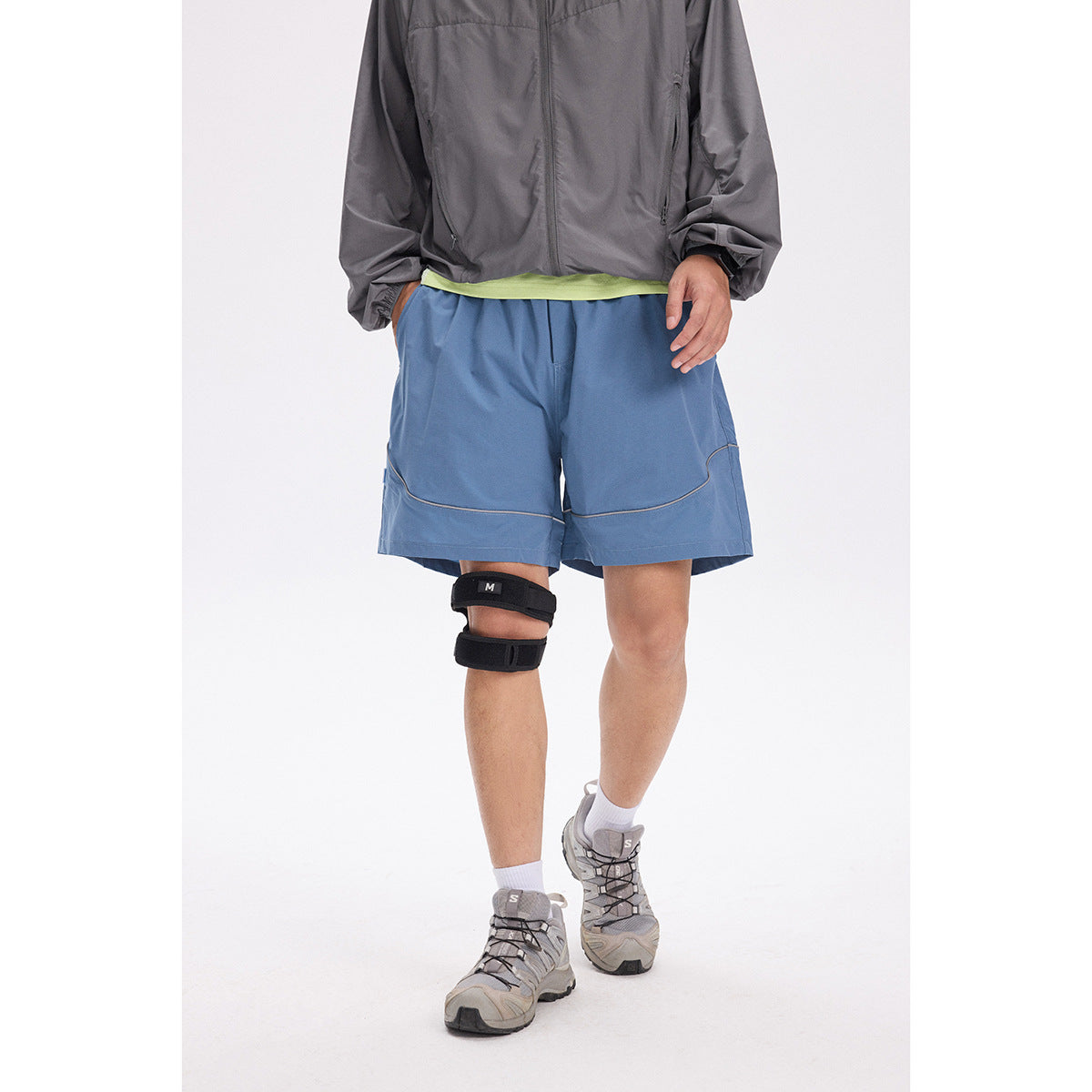 Model wearing blue athletic shorts with curved piping, a gray jacket and a lime shirt.