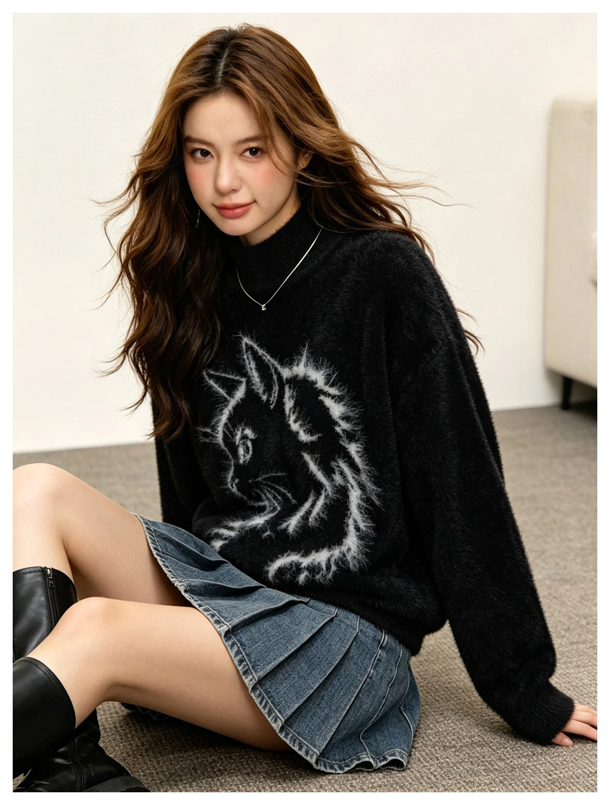 Woman sitting on a carpet wearing a black fuzzy cat-graphic sweater with a pleated denim mini skirt and black boots