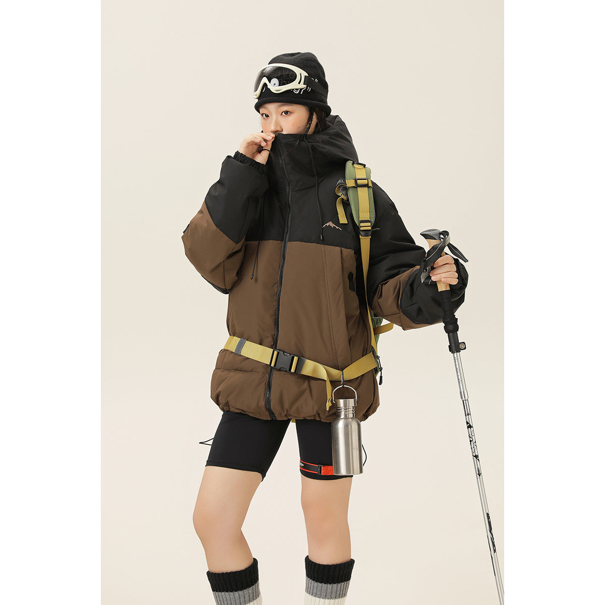 Model wearing a black-and-brown color-block hooded jacket with a yellow hiking backpack and trekking pole.