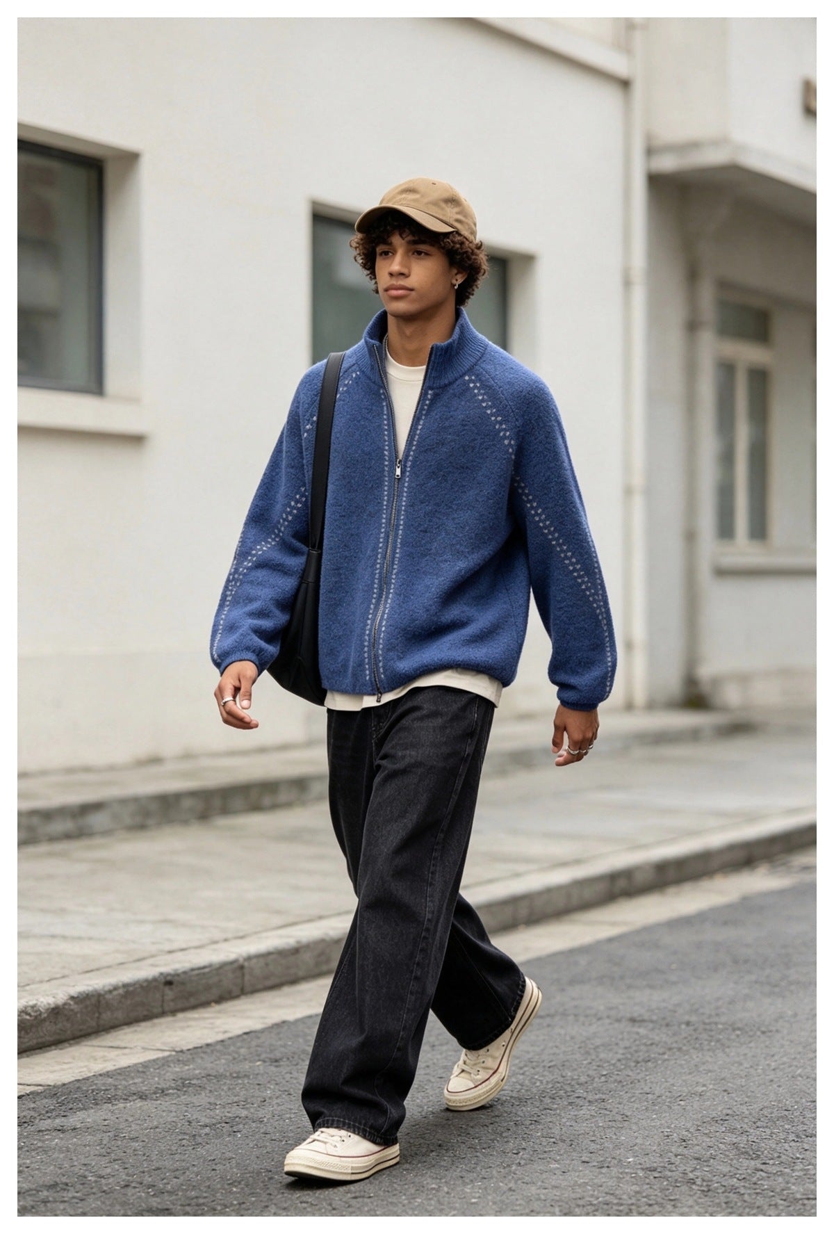 Man walking outdoors in a blue zip-up knit cardigan over a cream tee with black wide-leg jeans and a tan cap