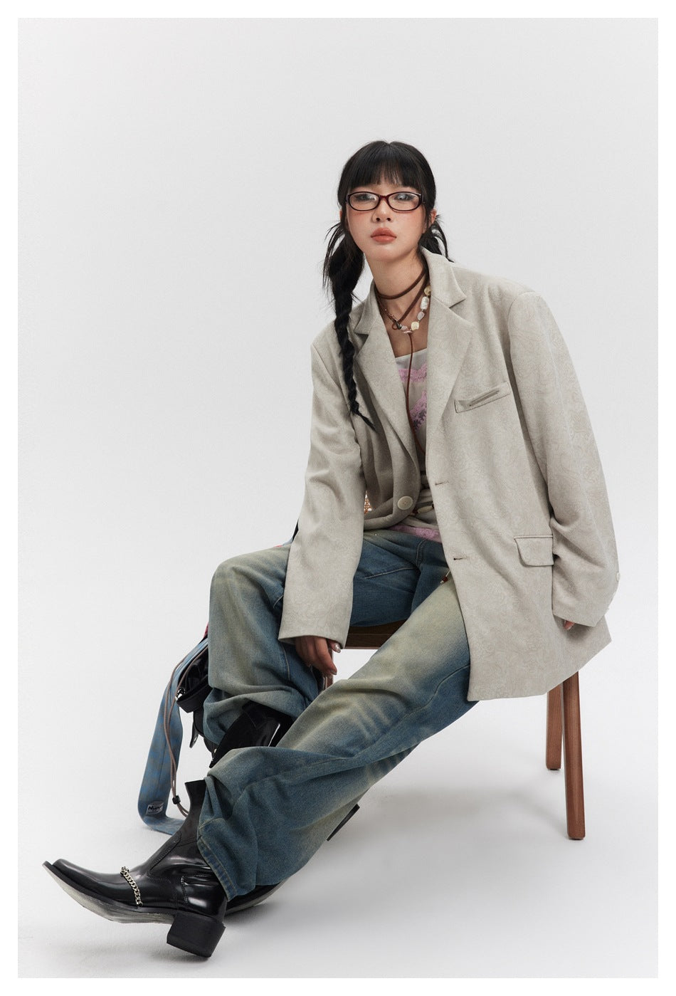 Model seated in a light beige patterned blazer with blue jeans and black boots.
