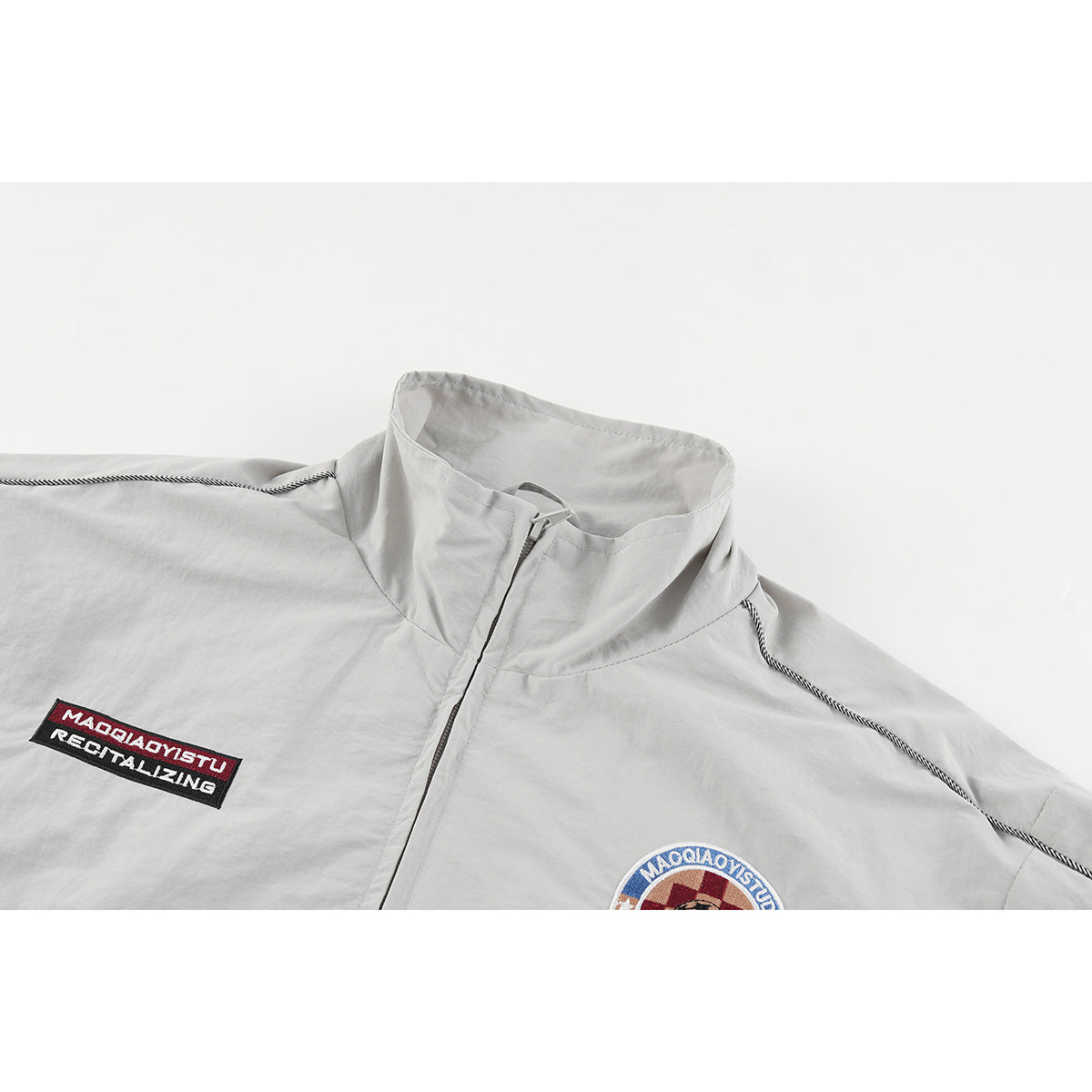 Close-up of a light gray zip-up jacket with a stand collar, contrast piping, and embroidered chest patches.